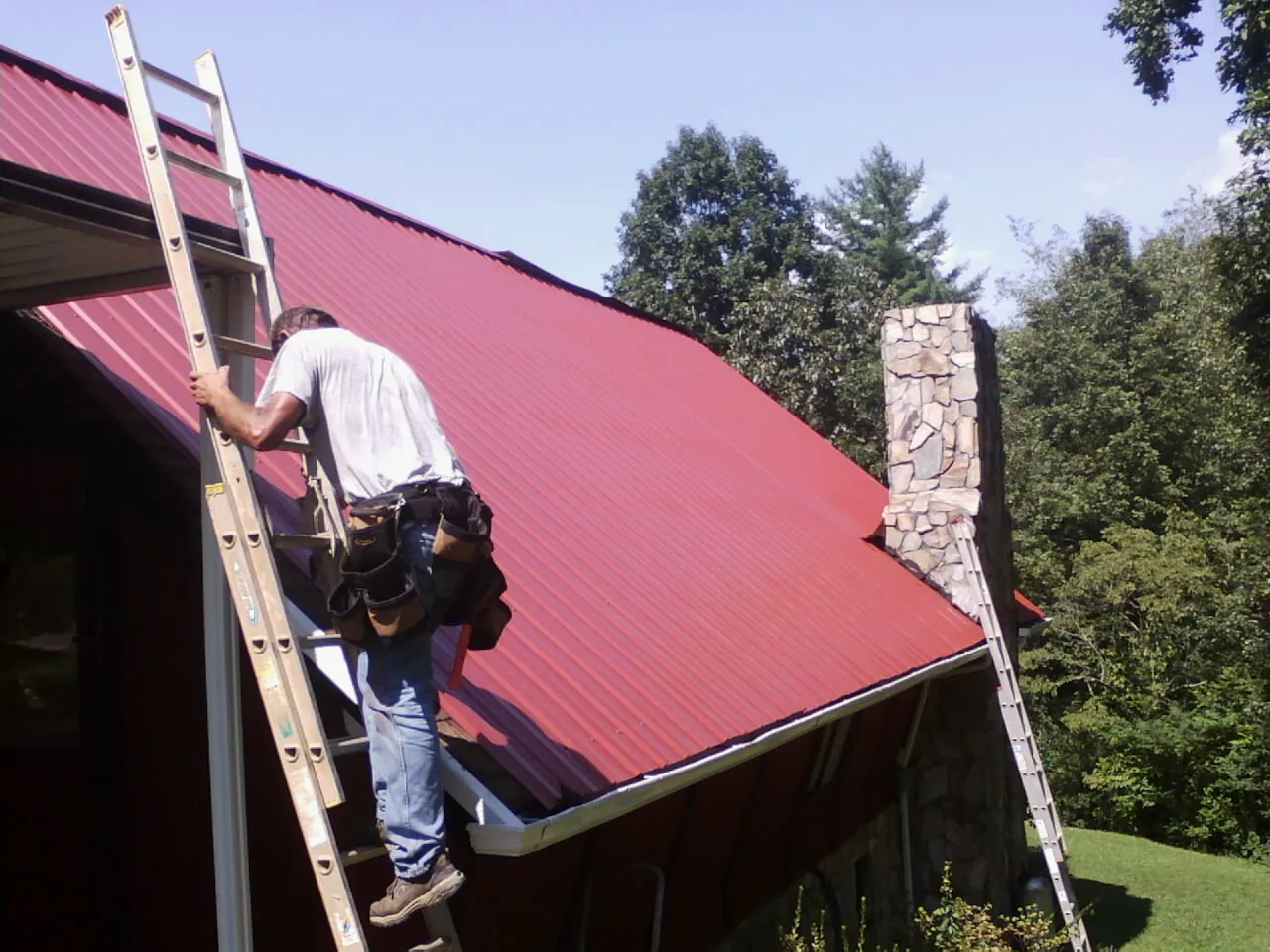 Roofer on a ladder beside a red metal roof and stone chimney — Soffit & Fascia Repair by Roofing Brothers in Grottoes